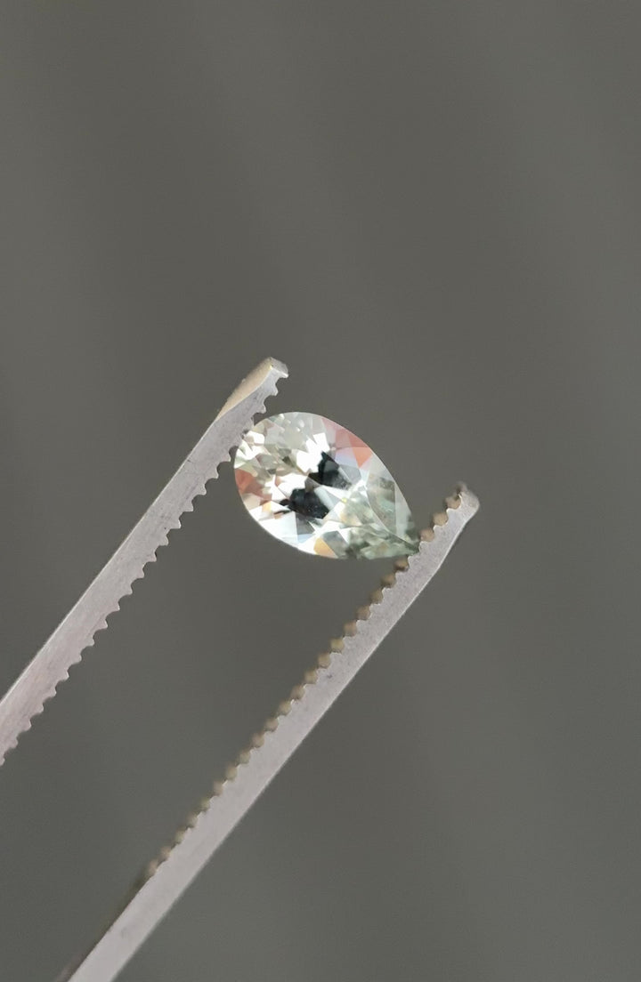 Tweezers-held video of a precision pear mixed cut Montana sapphire, rotating to reveal faint silver seafoam tones, clean facet junctions, and natural untreated sparkle in a loose gemstone.