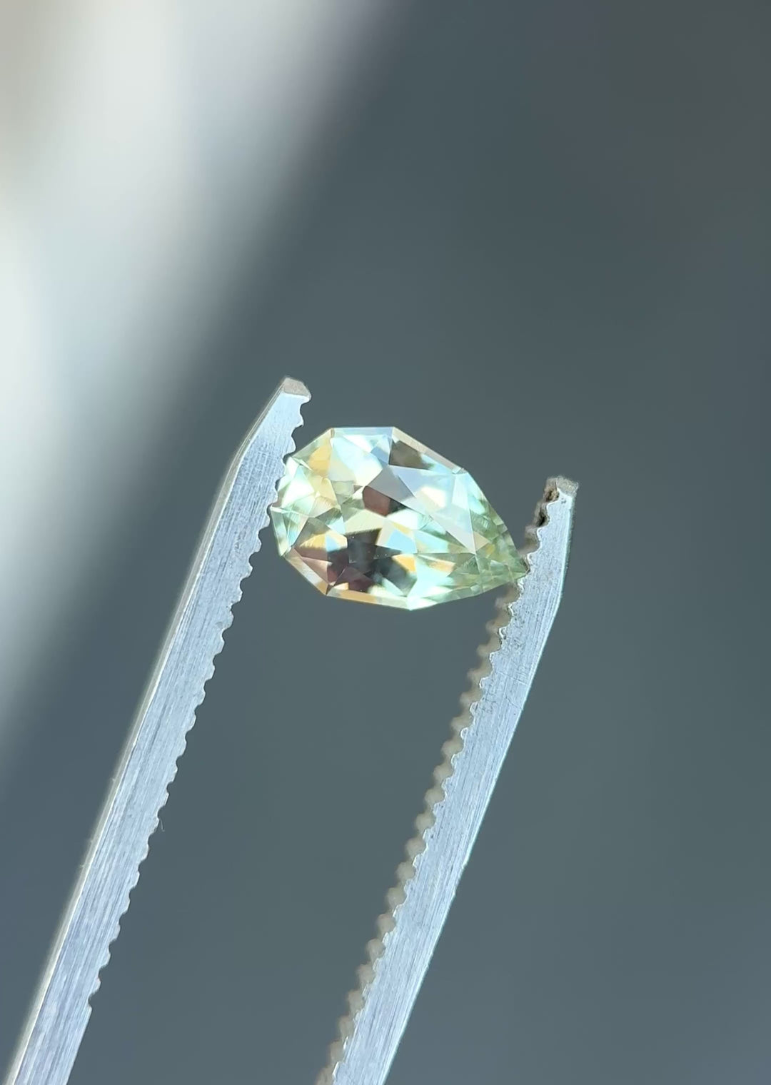 Natural Madagascar sapphire turning in tweezers, showcasing pear cut brilliance and subtle seafoam green color under changing light.