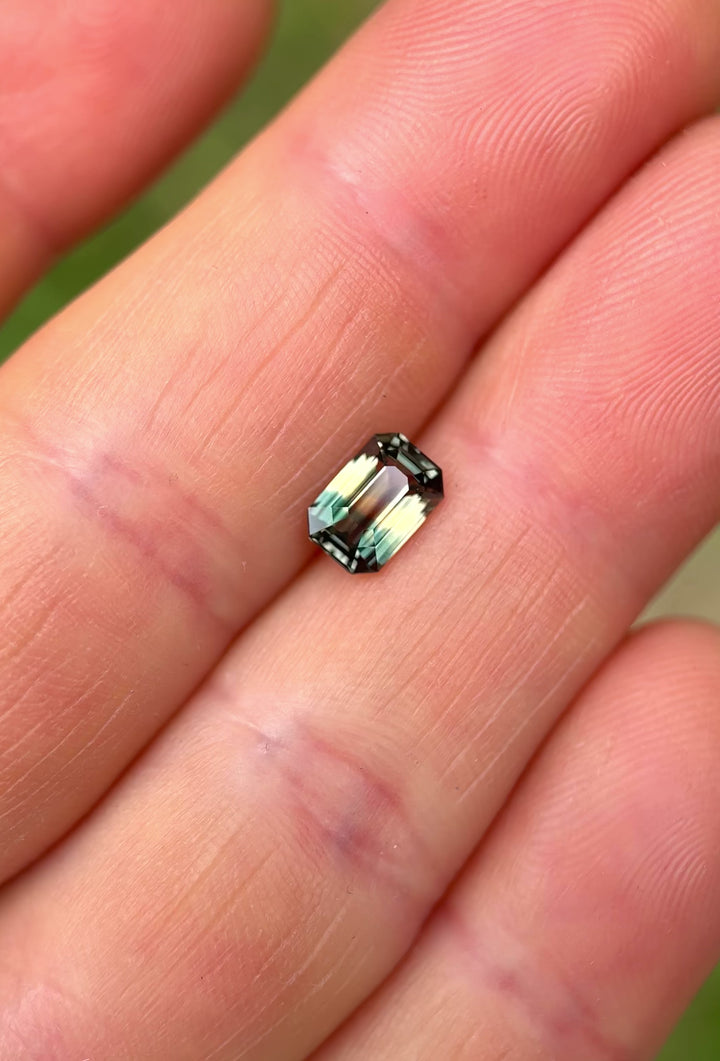 A 1.60ct precision emerald cut sapphire from Madagascar, showing strong fluorescence and a dynamic color shift from peach and green to mauve pink, held between fingers in natural light with a soft outdoor background.