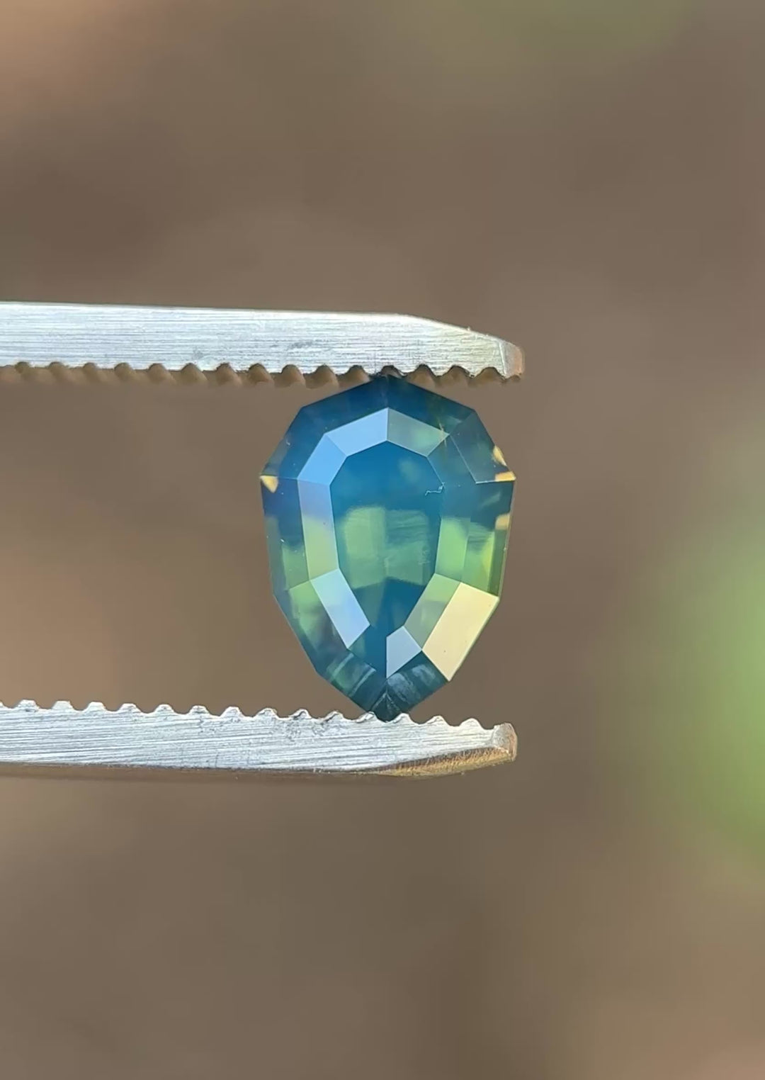 Natural unheated teal blue green opalescent sapphire held in tweezers, highlighting the geometric shield outline and reflective step-cut facets.