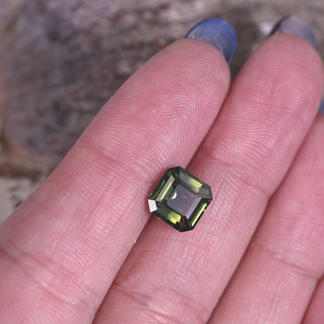 Loose 3.10 carat parti sapphire resting on a fingertip for scale, highlighting the square Asscher cut and the natural mix of green, yellow, and blue tones