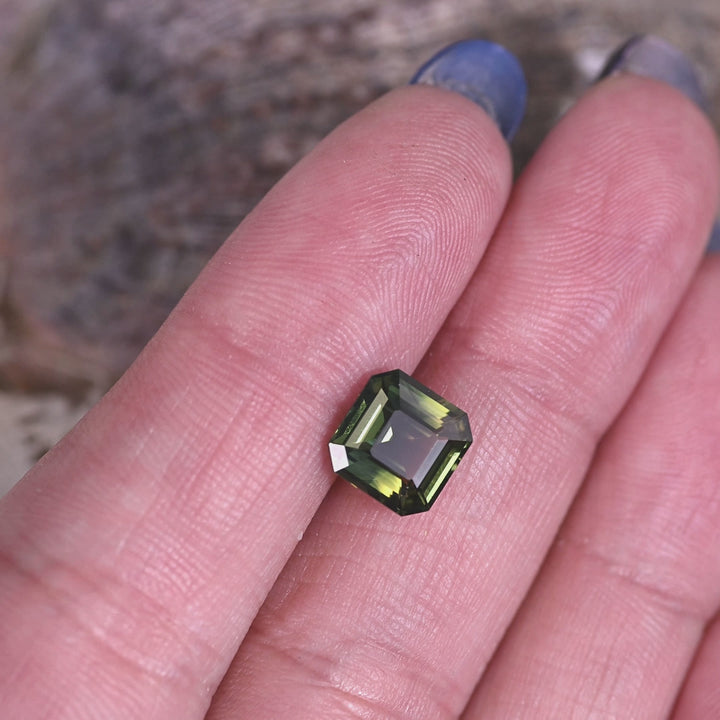 Loose 3.10 carat parti sapphire resting on a fingertip for scale, highlighting the square Asscher cut and the natural mix of green, yellow, and blue tones