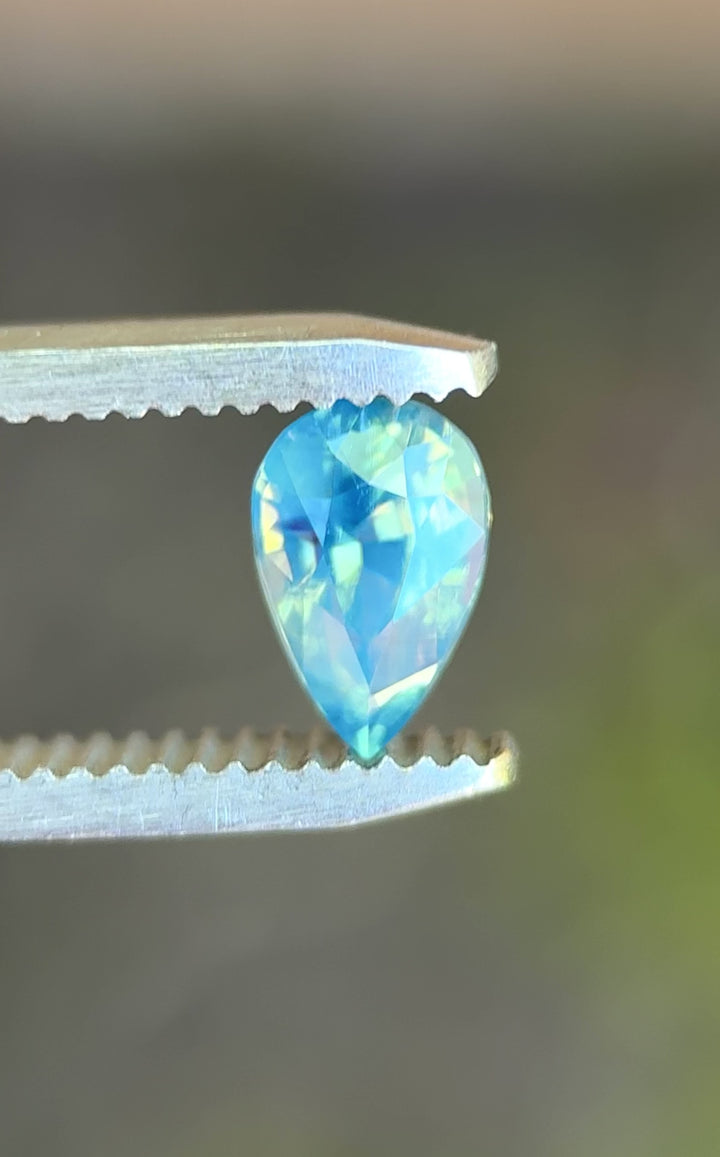 Natural Madagascar sapphire turning in tweezers, showcasing pear mixed cut symmetry and subtle teal-to-blue color movement under changing light.