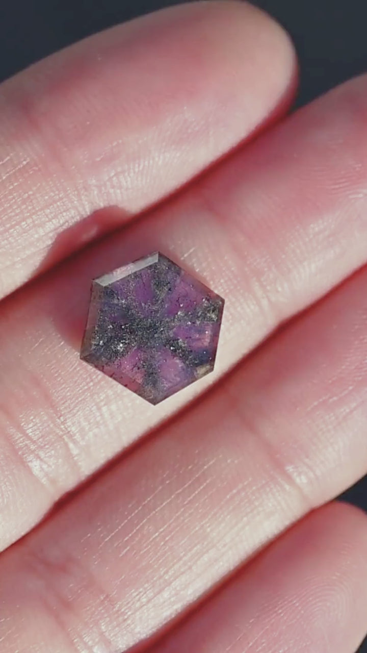 Explore a 5.56ct untreated pink purple sapphire from Afghanistan featuring a distinctive trapiche-like inclusion pattern. Displayed on the hand for scale, its broad hexagonal portrait cut reveals dark mineral inclusions radiating through translucent violet-pink crystal. An unusual collector's gemstone for bespoke jewelry, statement rings, and artistic pendants.