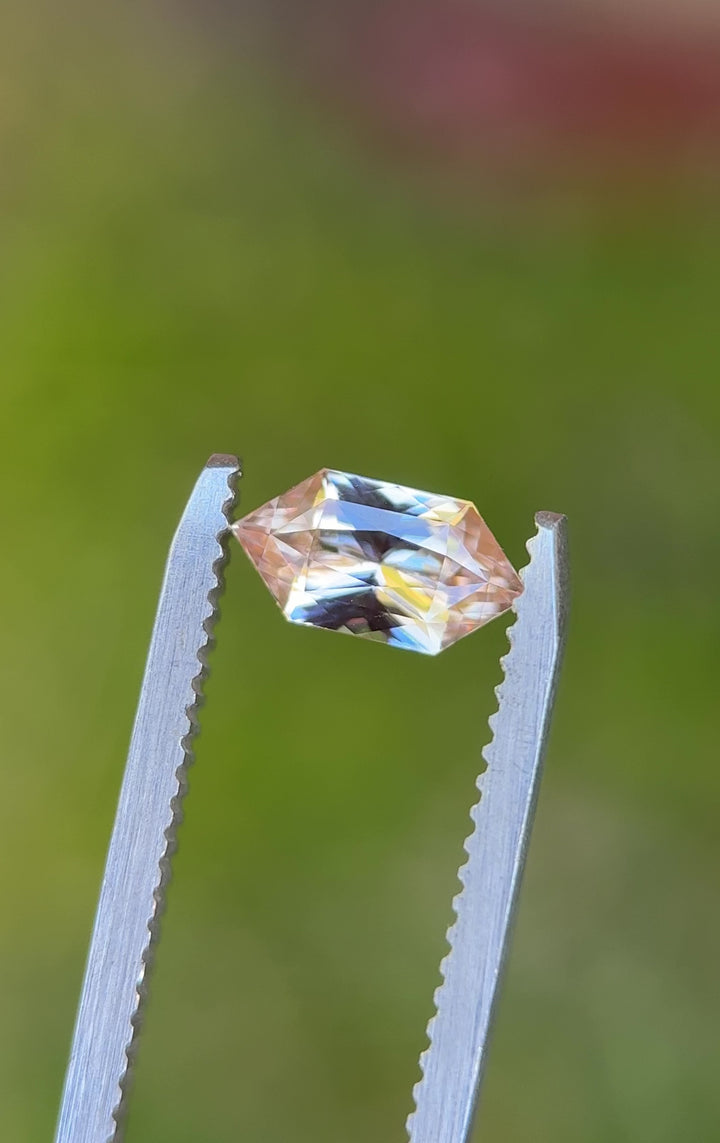 Elongated hexagon sapphire rotating in tweezers displaying custom modified brilliant facets and warm peach tones.