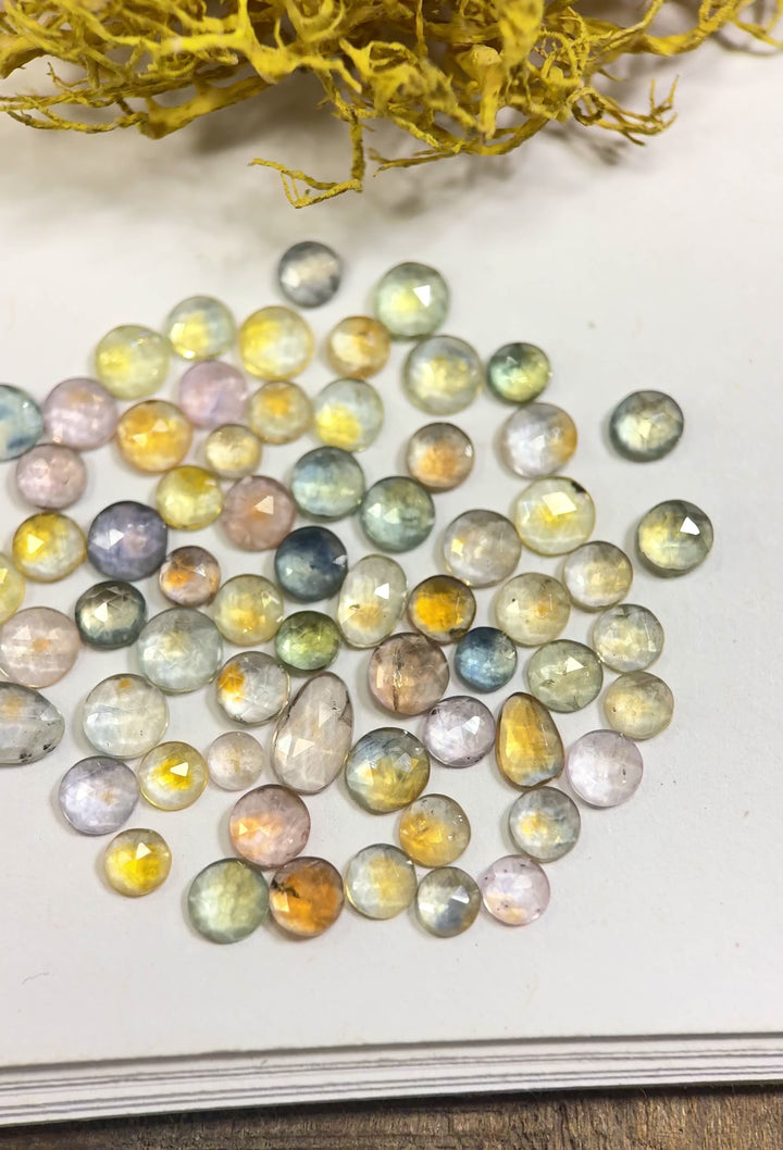 Video of 66 fancy parti sapphires from the Eldorado Deposit, Montana, glimmering under soft light to reveal their vivid teal, yellow, orange, and pink hues. The 17.40-carat parcel includes mixed round, oval, and freeform cuts, all heat-treated for vibrant color.