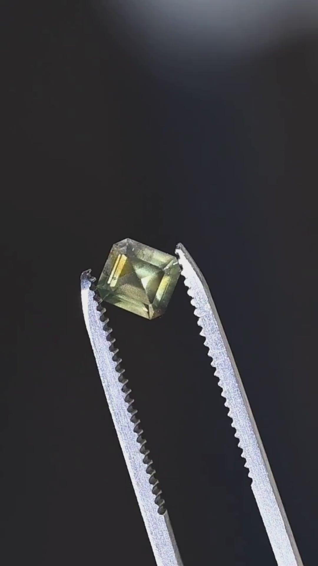 Explore the geometric faceting of this untreated 1.08ct opalescent moss green sapphire from Nigeria. Held in jeweler's tweezers, the gemstone features a precision octagonal step mix cut, olive-green body color and golden-green reflections.