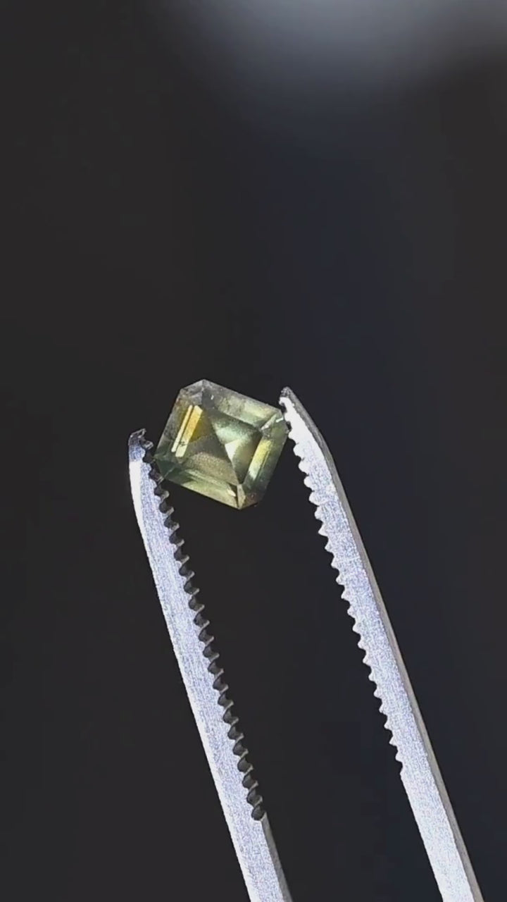 Explore the geometric faceting of this untreated 1.08ct opalescent moss green sapphire from Nigeria. Held in jeweler's tweezers, the gemstone features a precision octagonal step mix cut, olive-green body color and golden-green reflections.
