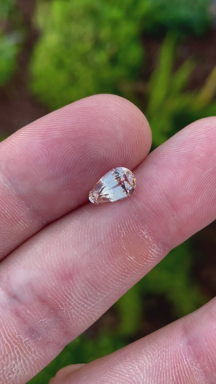 A close-up view of a 2.45-carat sapphire in pastel champagne and peach hues. The video focuses on the brilliant sparkle as the stone moves slightly, allowing viewers to appreciate its custom teardrop cut and the play of light across its facets. The gemstone is untreated, sourced from Sri Lanka, and measures 10.12mm in length, 6.40mm in width, and 4.70mm in height.