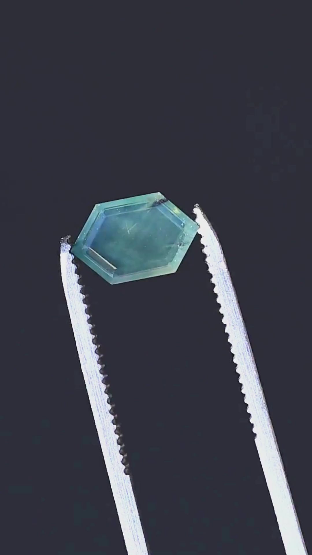 A close-up presentation of a 2.15ct untreated opalescent teal green sapphire from Gombe, Nigeria. Held in jeweler's tweezers, the gemstone features an elongated hexagonal portrait cut, a broad translucent central window and fine internal striations.