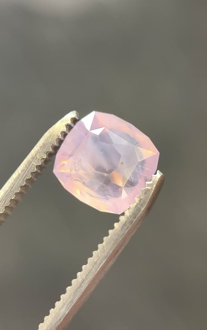 Ballet pink opalescent sapphire rotating in gem tweezers showing off whispy inclusions and natural opalescence.