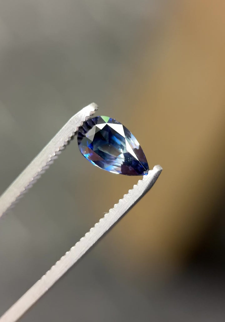 Video of 2.02CT bi-color blue sapphire rotating in gemstone tweezers under natural light, revealing vivid color shifts and scintillation from the pear brilliant cut, natural unheated sapphire from Sakaraha, Madagascar.