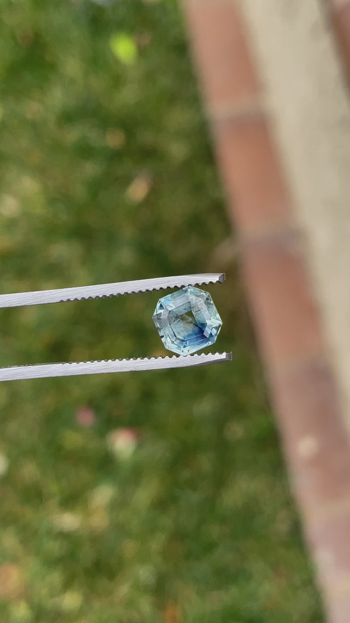 A rotating video of a 2.45-carat parti blue-green sapphire in an antique emerald cut, held by tweezers against a soft background, highlighting its symmetry and vivid colors.