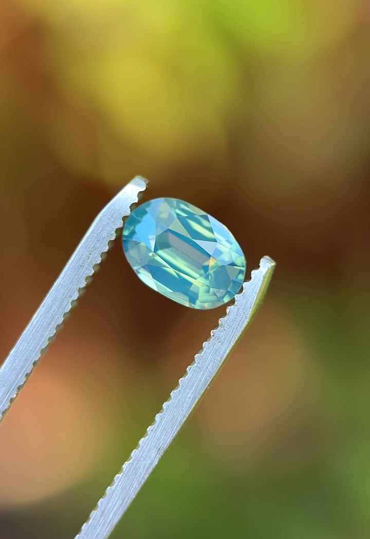 Natural Madagascar sapphire turning in tweezers, showcasing elongated oval symmetry and subtle teal-to-blue color movement under changing light.