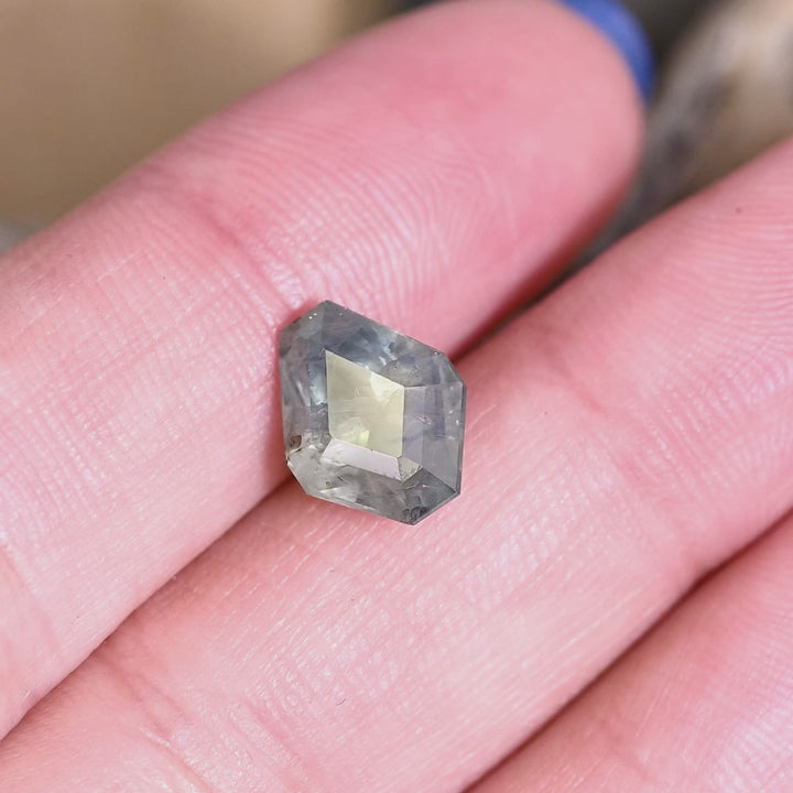 3.78ct teal and blue Montana sapphire in geometric tablet kite cut rotating slowly on fingertips, showing color shift from deep inky navy at the center to brighter sea-teal flashes along the angular upper facets, with visible internal silk and natural inclusions catching the light as the stone tilts.
