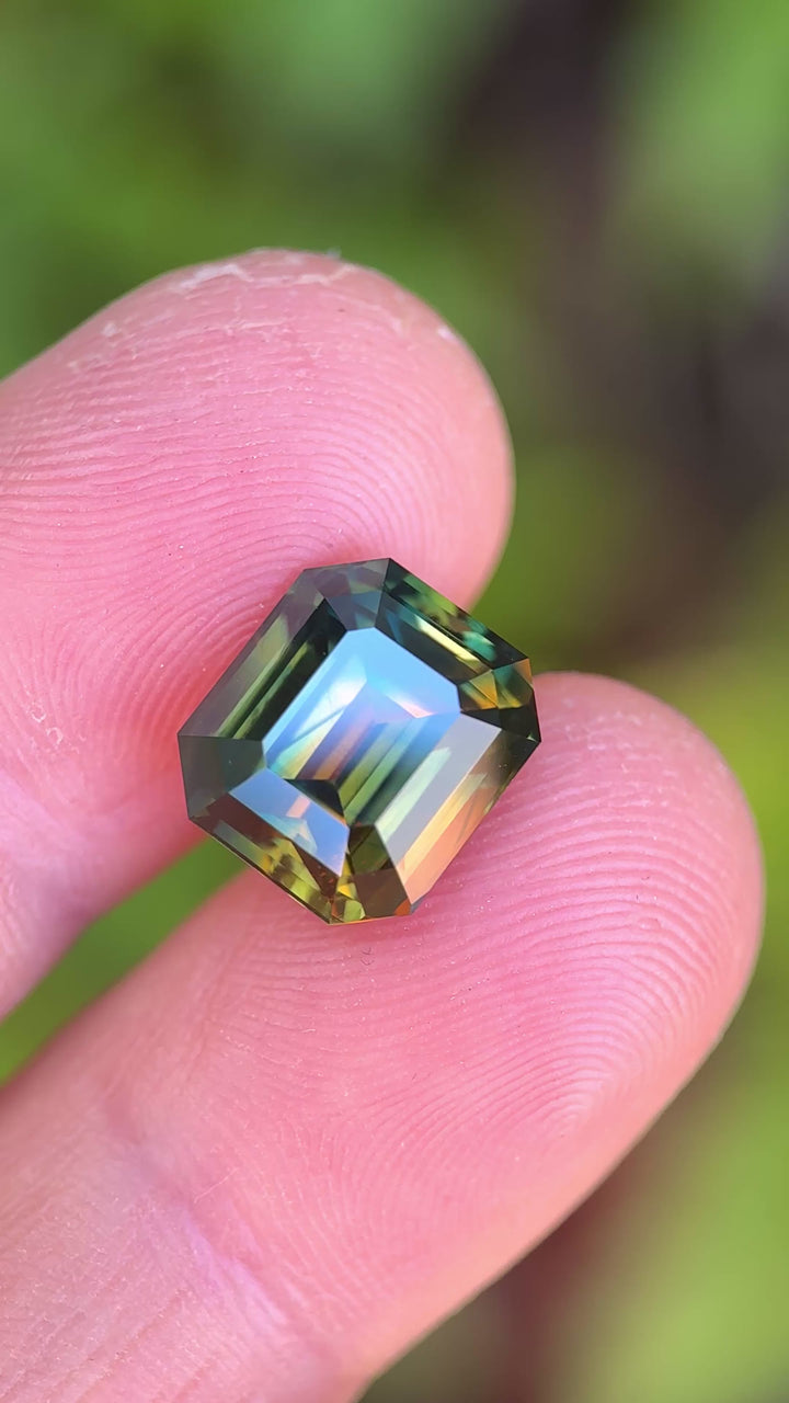 Video of a 9.14 carat Nigerian parti sapphire in a precision emerald cut, rotating to reveal orange, yellow, blue, and green color zoning with high clarity and depth, Gombe origin