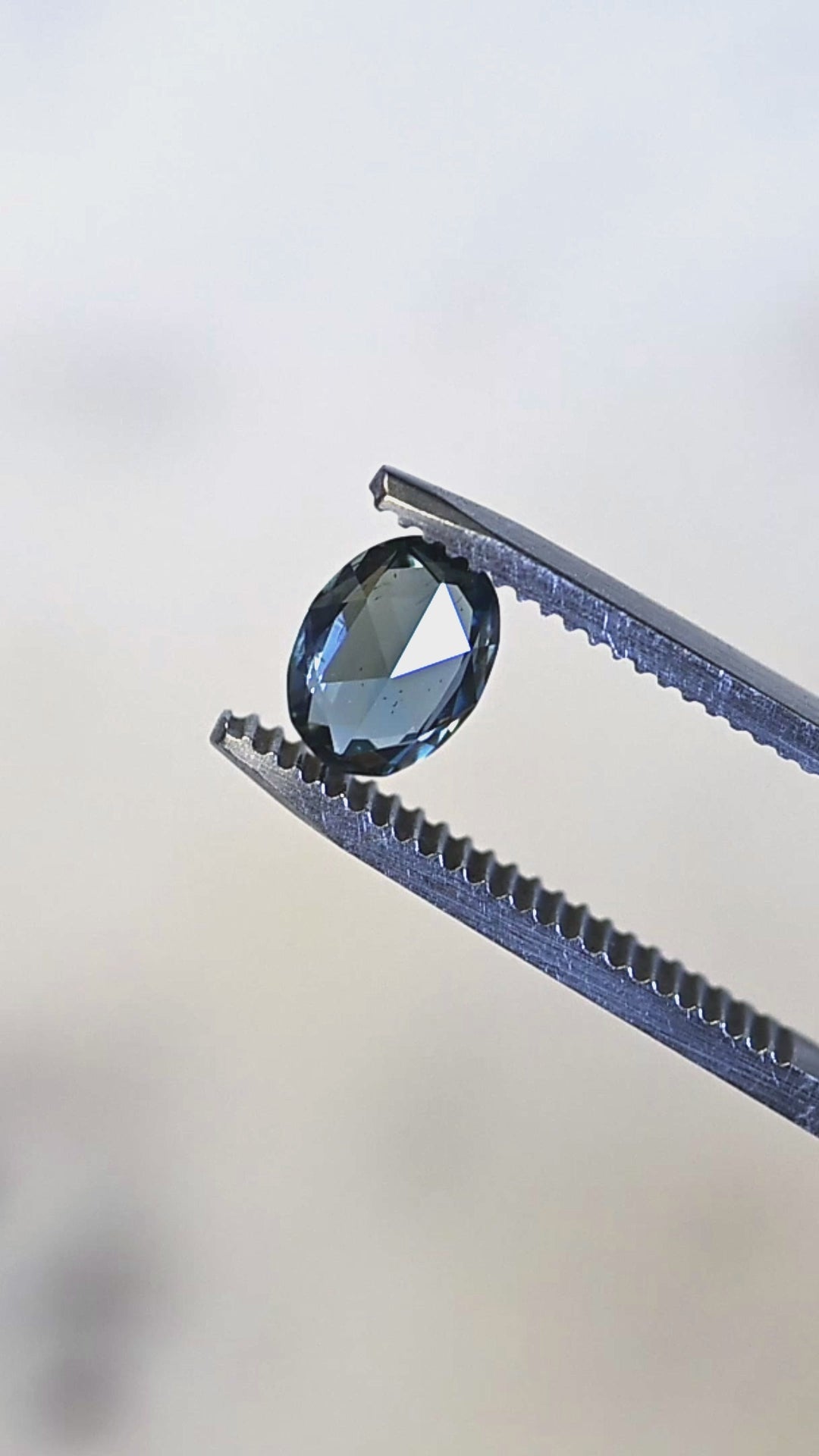 Macro video of a 1.37ct untreated Madagascar sapphire turning in tweezers, showcasing teal, blue, and green flashes across the faceted crown.