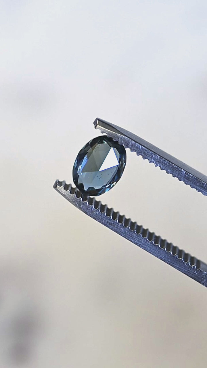 Macro video of a 1.37ct untreated Madagascar sapphire turning in tweezers, showcasing teal, blue, and green flashes across the faceted crown.