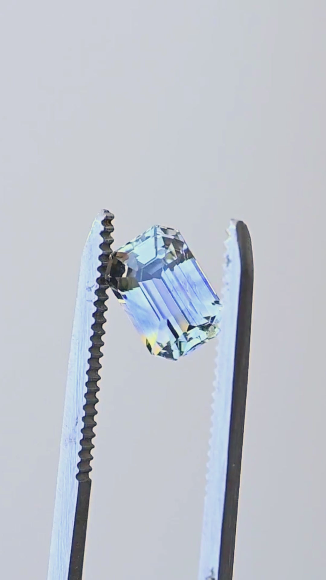 Precision emerald-cut Sri Lankan parti sapphire rotating in jeweler’s tweezers, showing its natural blue and yellow zoning, clipped corners and broad reflective step facets.