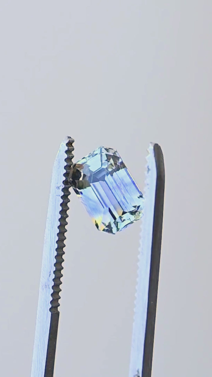 Precision emerald-cut Sri Lankan parti sapphire rotating in jeweler’s tweezers, showing its natural blue and yellow zoning, clipped corners and broad reflective step facets.