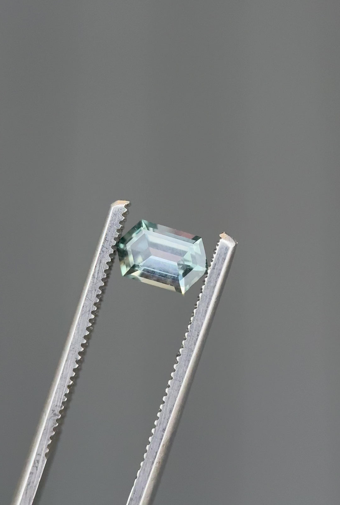 Close-up video of an unheated Montana sapphire turning in tweezers, demonstrating elongated hexagon step cut symmetry and visible color shift from seafoam to purple tones