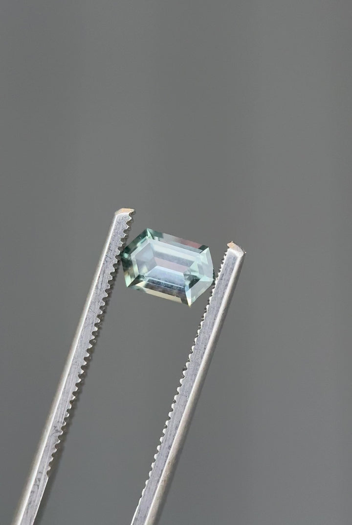 Close-up video of an unheated Montana sapphire turning in tweezers, demonstrating elongated hexagon step cut symmetry and visible color shift from seafoam to purple tones