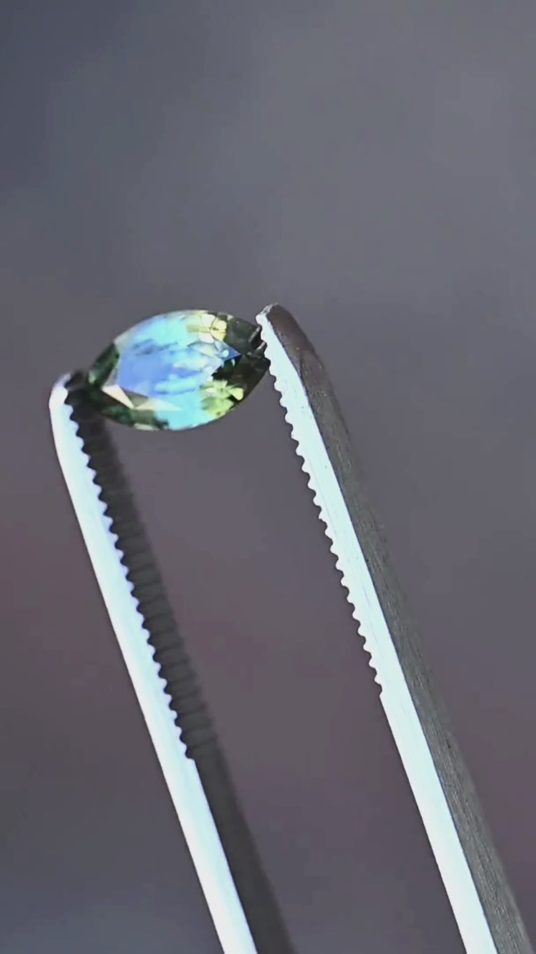 A close-up presentation of a 2.03ct teal blue yellow parti sapphire from Diego, Madagascar. Held in jeweler's tweezers, the gemstone displays contrasting teal-blue, olive-green and golden-yellow reflections through its custom faceting.