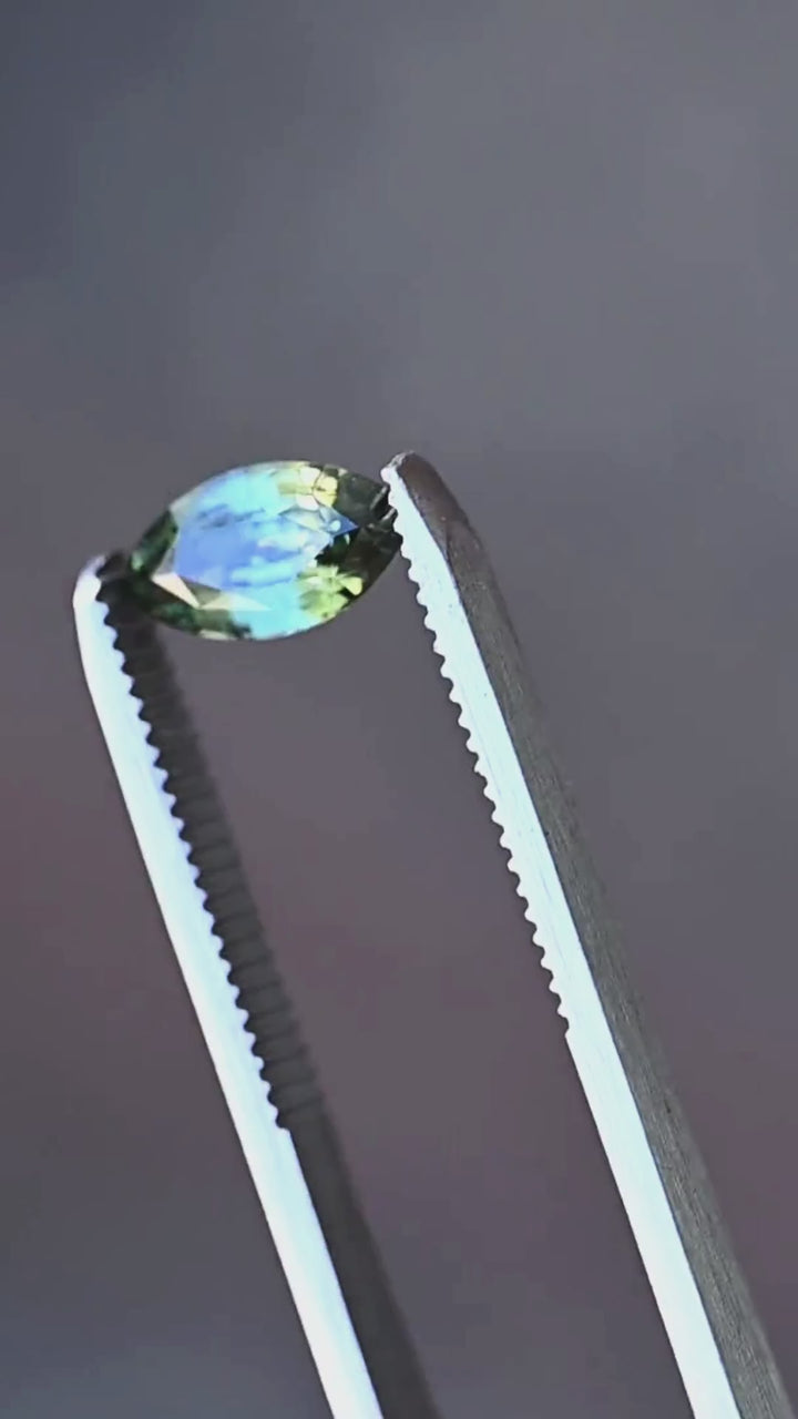 A close-up presentation of a 2.03ct teal blue yellow parti sapphire from Diego, Madagascar. Held in jeweler's tweezers, the gemstone displays contrasting teal-blue, olive-green and golden-yellow reflections through its custom faceting.