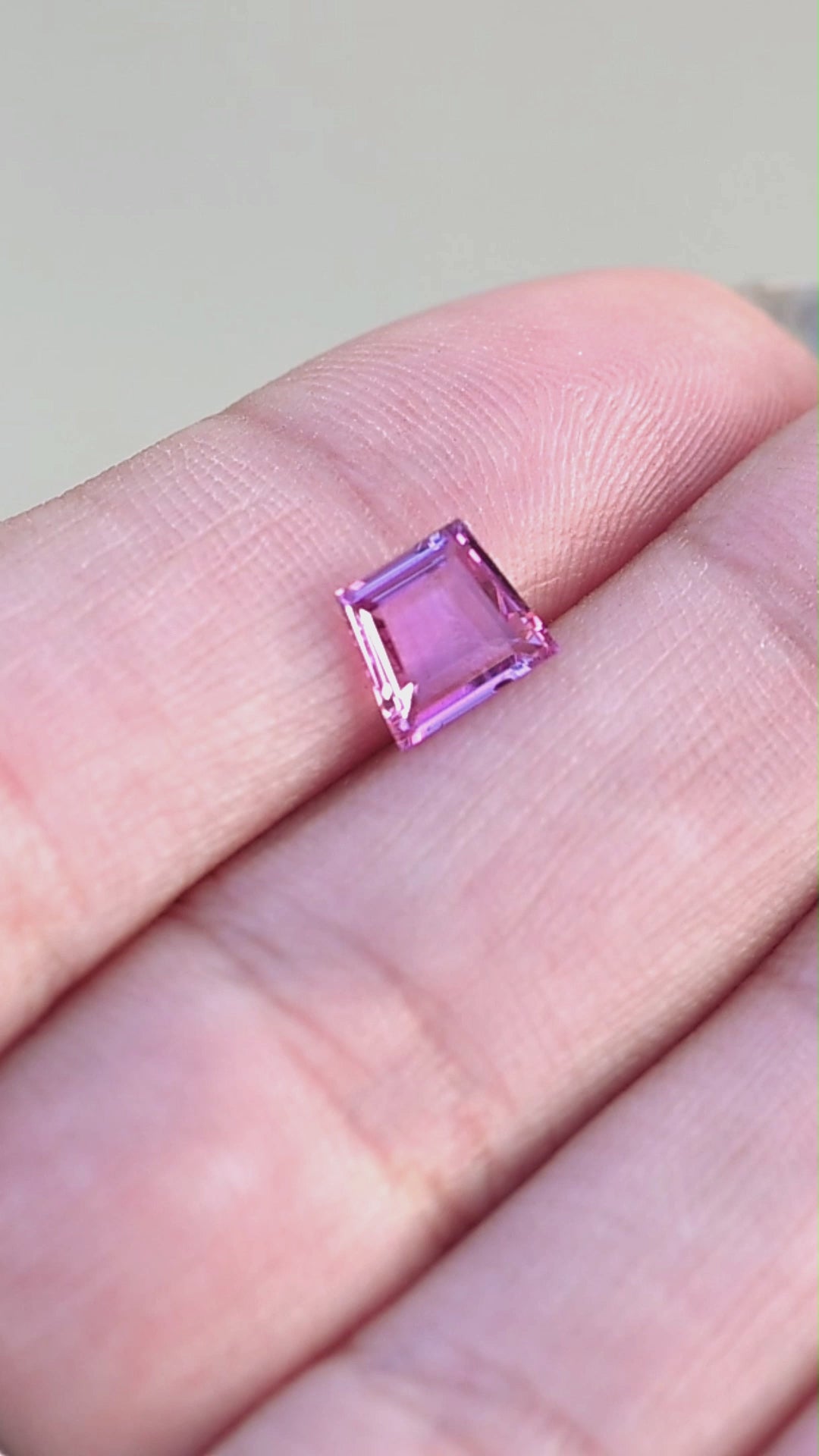 Hot pink natural sapphire in a trapezoid shaped portrait cut held in fingertips rotating under open shade.