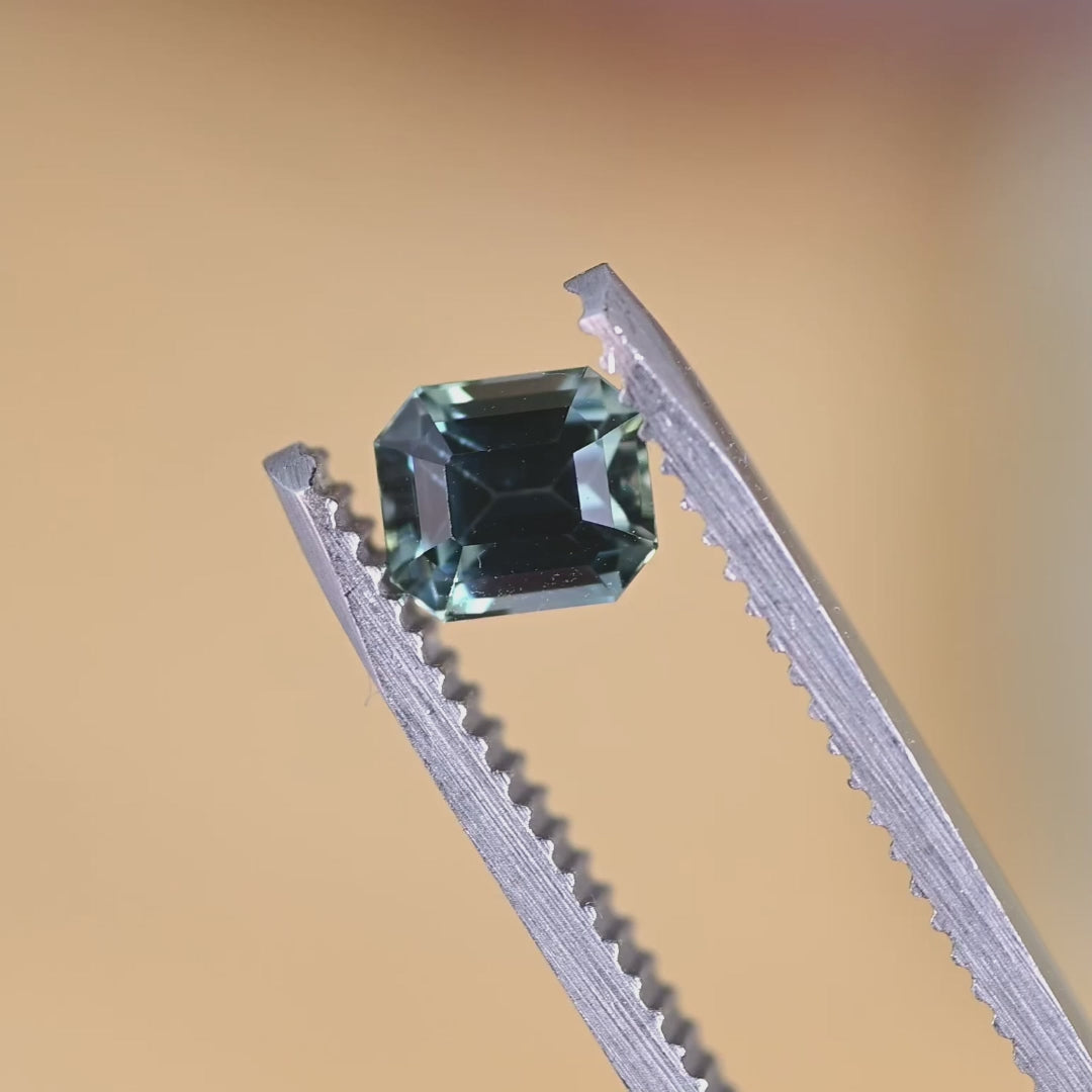 Loose emerald cut sapphire turning in tweezers, revealing natural blue and green color shifts from Sakaraha, Madagascar.