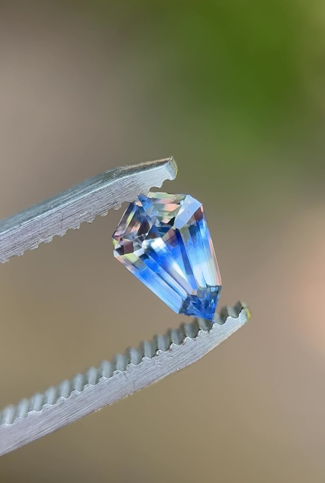 Rotating shield-cut 1.04ct Parti sapphire from Sri Lanka held in tweezers, displaying blue and colorless flashes under natural light.