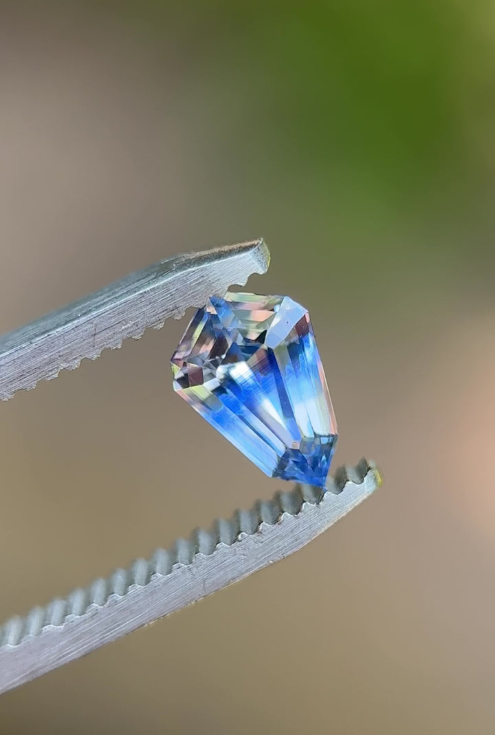 Rotating shield-cut 1.04ct Parti sapphire from Sri Lanka held in tweezers, displaying blue and colorless flashes under natural light.
