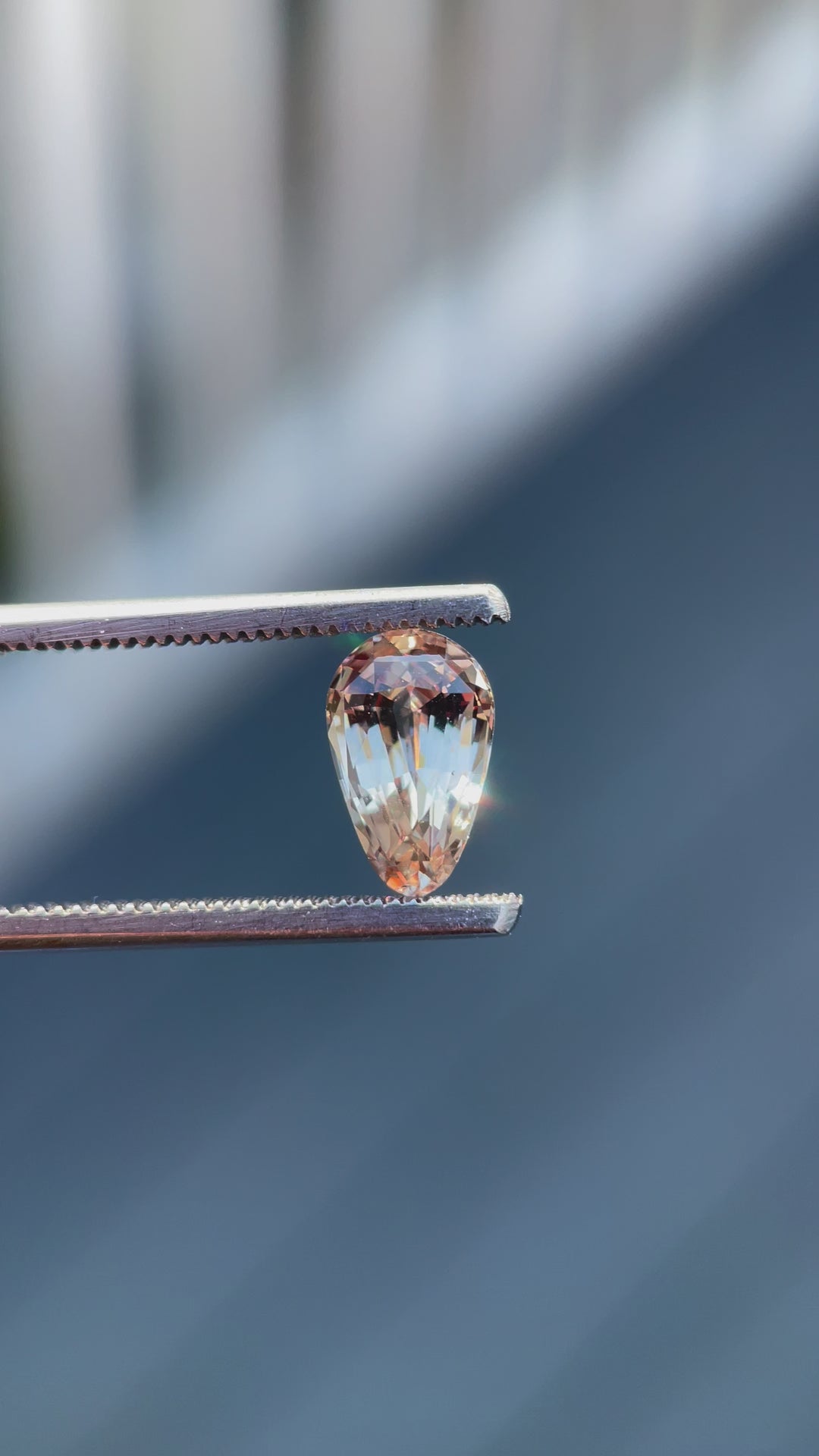 A 2.45-carat pastel champagne and peach sapphire is being rotated slowly under natural sunlight to showcase its custom brilliant teardrop cut. The light captures its intricate facets and the delicate play of colors, highlighting the gemstone's clarity and luster. Originating from Sri Lanka, this untreated sapphire measures 10.12mm in length, 6.40mm in width, and 4.70mm in height.