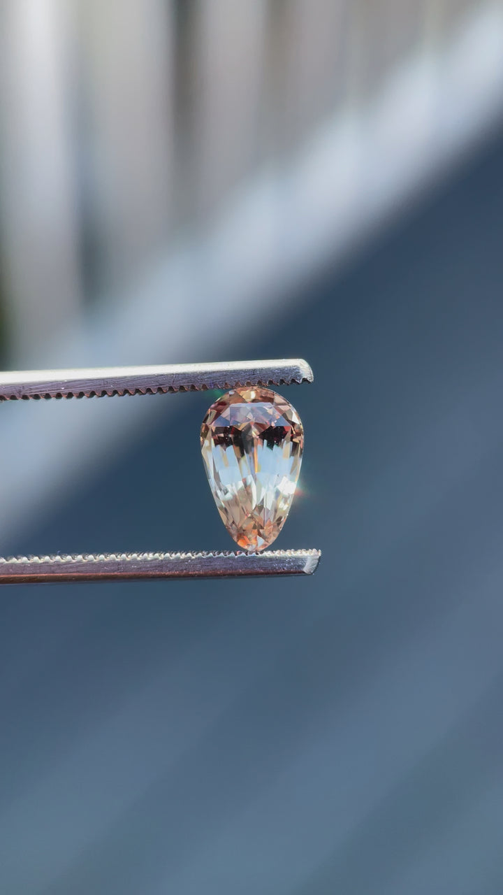 A 2.45-carat pastel champagne and peach sapphire is being rotated slowly under natural sunlight to showcase its custom brilliant teardrop cut. The light captures its intricate facets and the delicate play of colors, highlighting the gemstone's clarity and luster. Originating from Sri Lanka, this untreated sapphire measures 10.12mm in length, 6.40mm in width, and 4.70mm in height.
