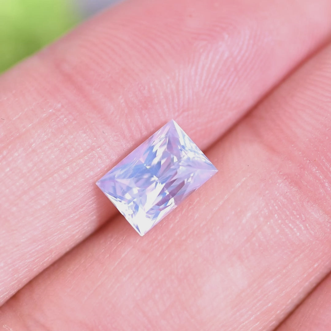 Rotating loose opalescent white sapphire from Ratnapura, Sri Lanka shown on the hand, revealing soft lilac flashes and bright reflections from the elongated princess cut.