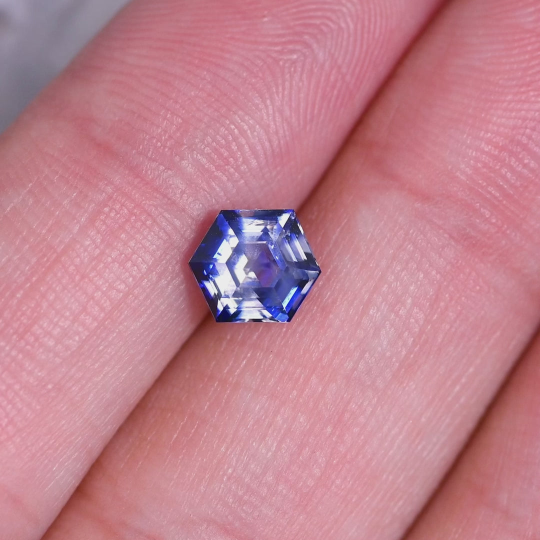 Rotating loose bi‑color blue and colorless sapphire from Ratnapura, Sri Lanka shown in hand, revealing the geometric hexagon step cut and changing reflections as it moves in light.