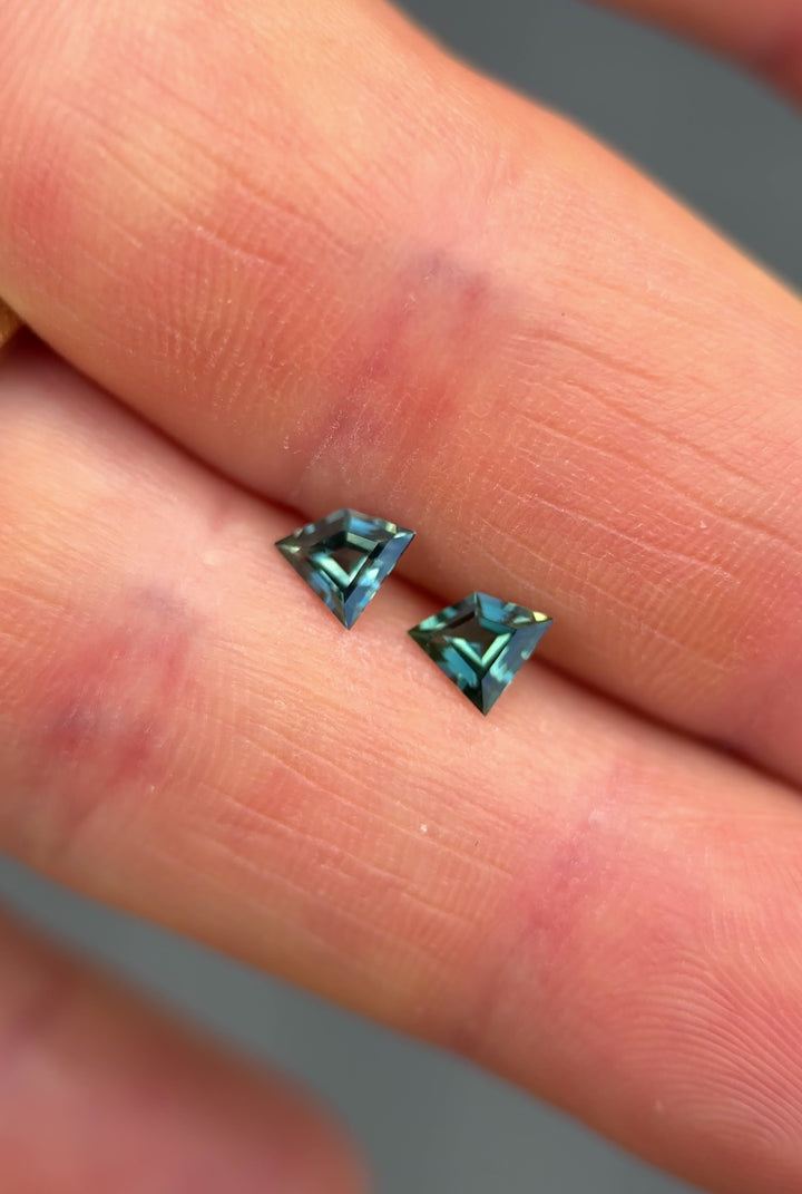 Video of a pair of teal blue-green kite step cut sapphires rotating in hand under natural light, showing color change, brilliance, and clean facets. Unheated 0.84ct sapphire pair from Queensland Gemfields, Australia.