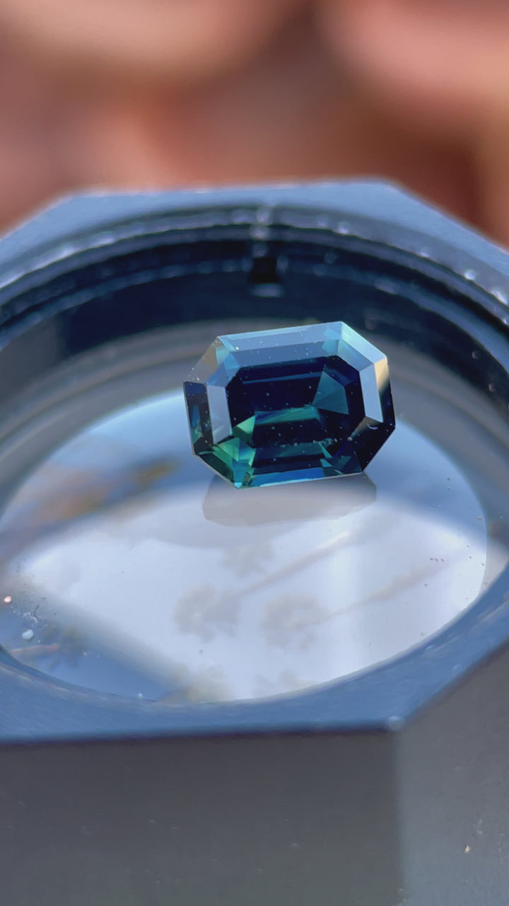 Video of a stunning step-cut sapphire resting on a metallic surface, reflecting the unique green and blue hues of this ethically sourced 1.16-carat gem.