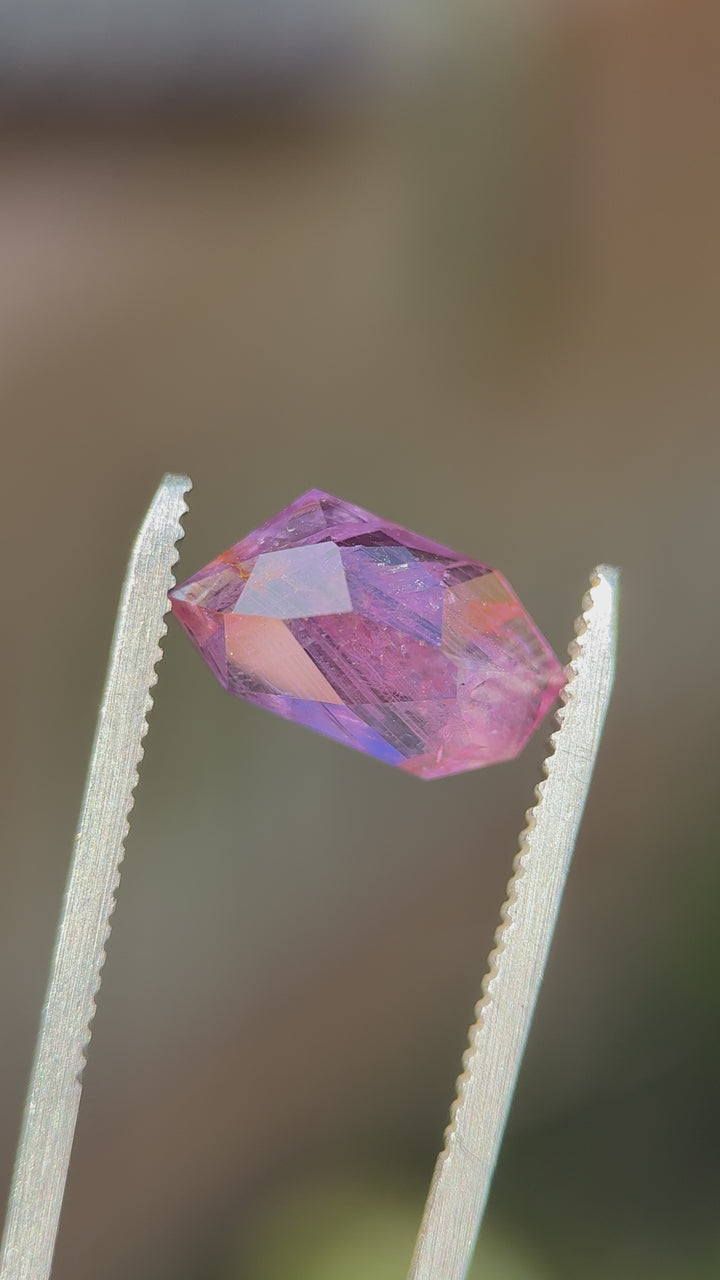Close-up of elongated pink-purple sapphire outdoors, emphasizing internal feathering and color depth across the angled crown facets.