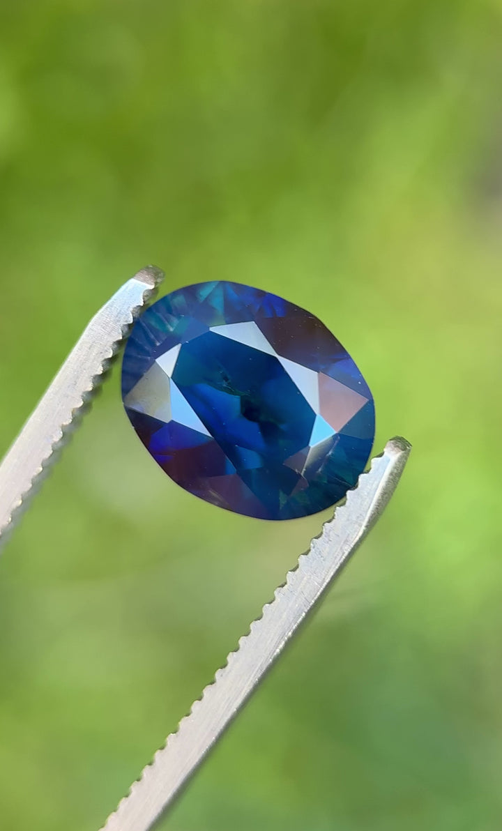Deep teal sapphire turned in tweezers under outdoor light, showing modified brilliant faceting and internal silk texture.