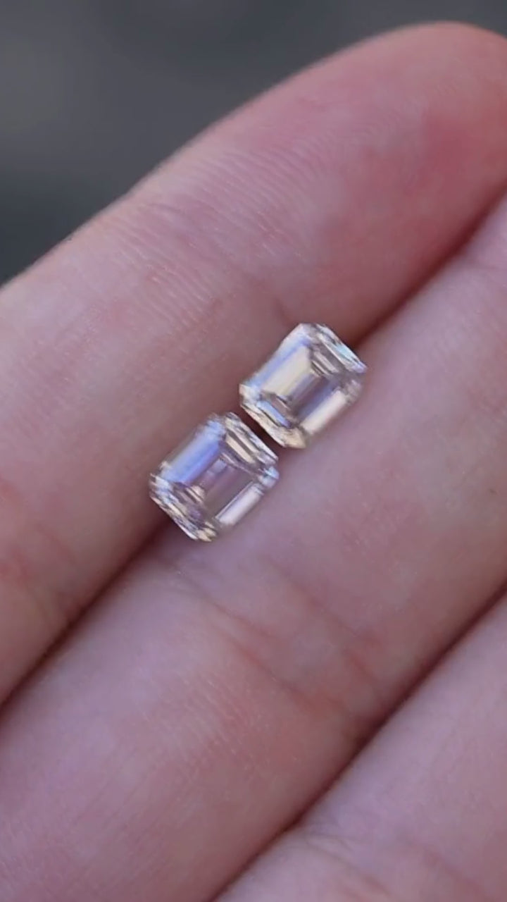 Matched 2.13 carat total weight emerald cut Canadian diamond pair moving in shaded natural light, revealing cool silver, blush and muted rosy-brown reflections across their broad step facets.