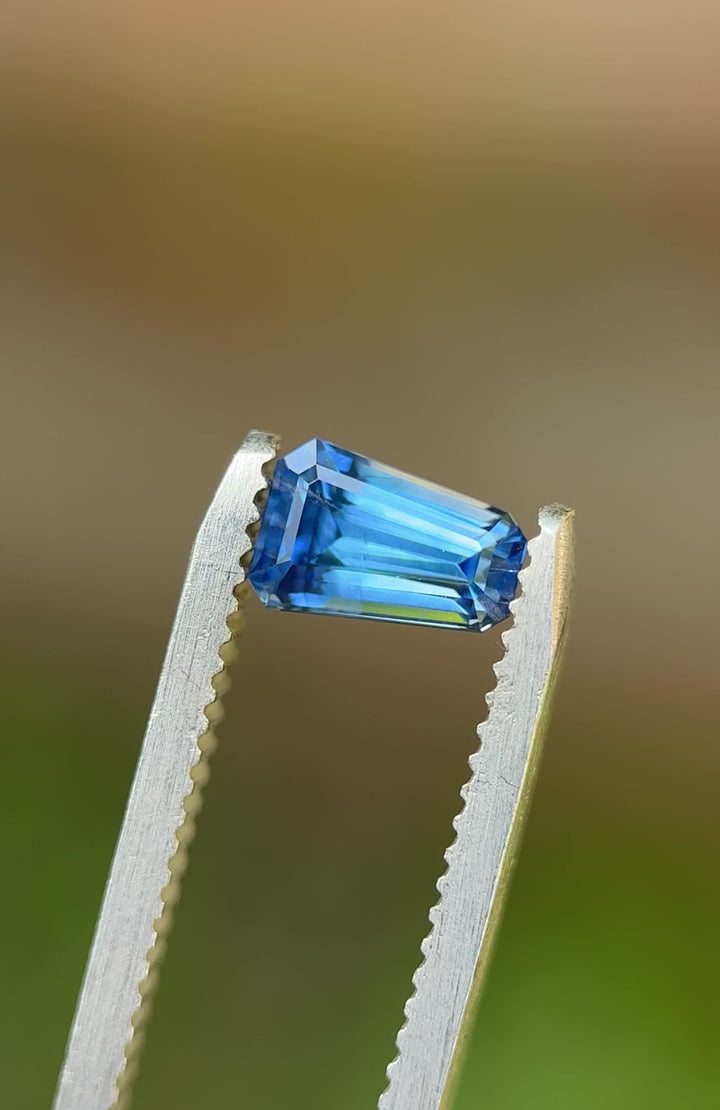 Precision-cut 1.20CT Montana sapphire in tweezers, rotated under sunlight to showcase vibrant blue color and symmetrical step-cut facets.