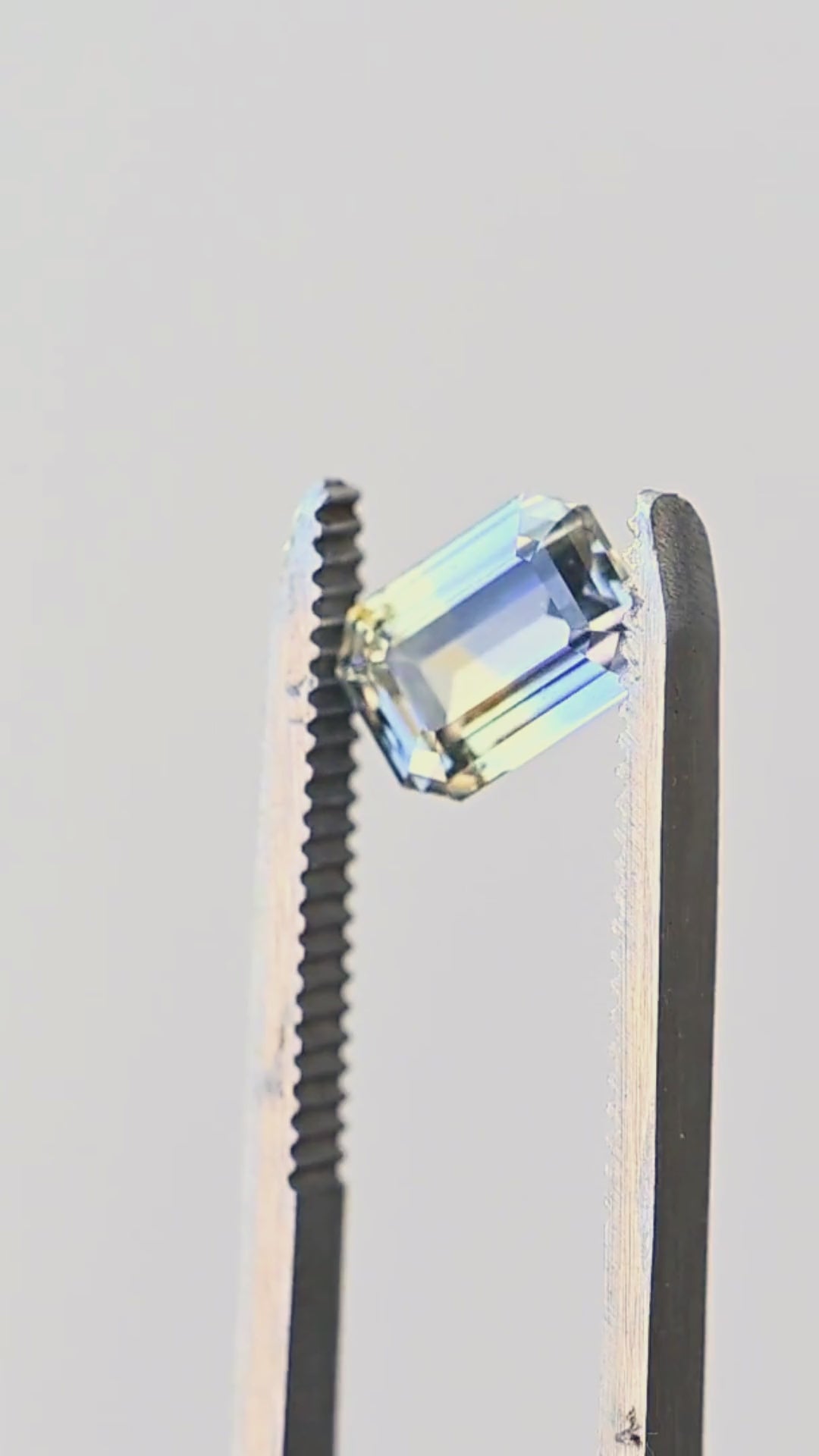 Blue and yellow parti sapphire rotating in jeweler’s tweezers, showing the crisp geometry, clipped corners and broad step facets of its precision custom emerald cut.