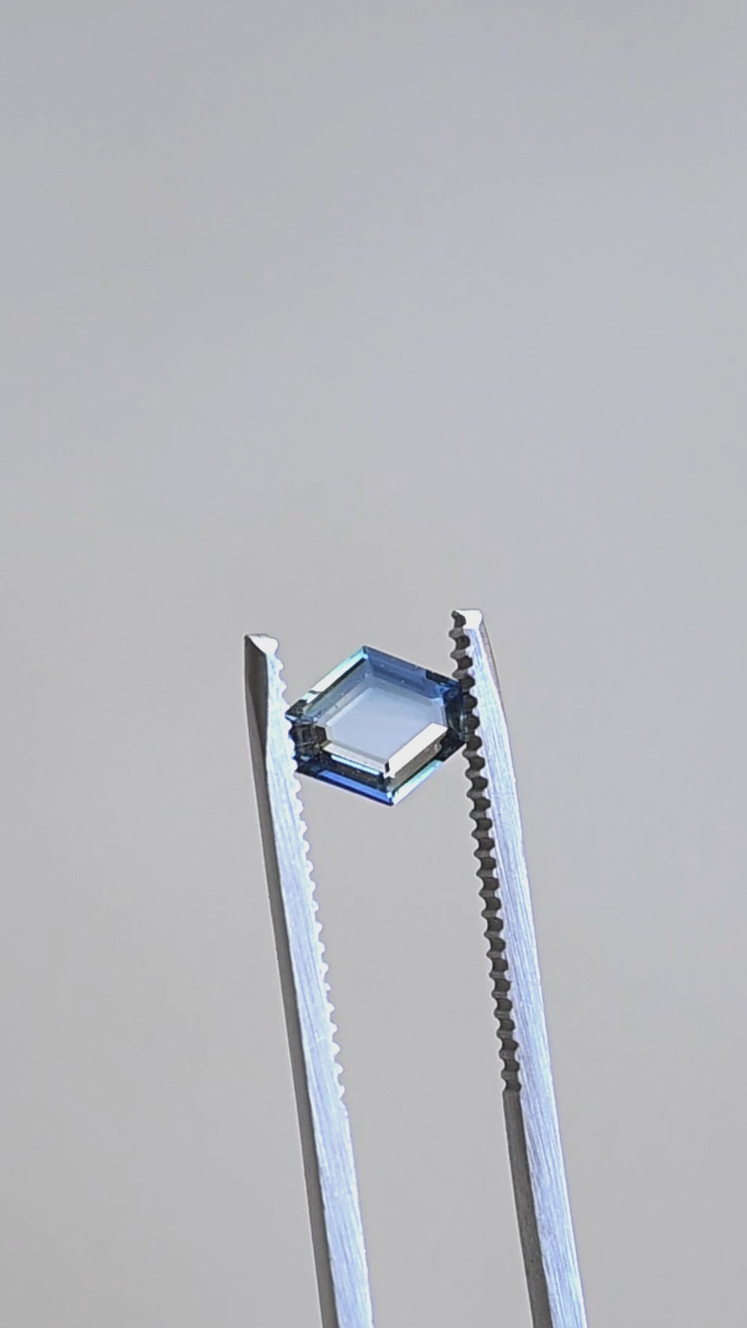Hexagonal portrait cut teal blue 1.02ct sapphire held in gem tweezers rotating under natural light.