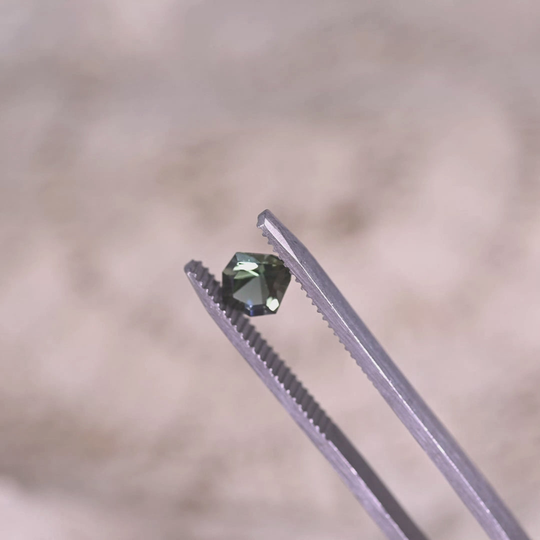 Loose teal blue green tanzanite from Merelani Hills Tanzania rotating in tweezers showing the geometric shield cut and shifting teal and blue reflections.