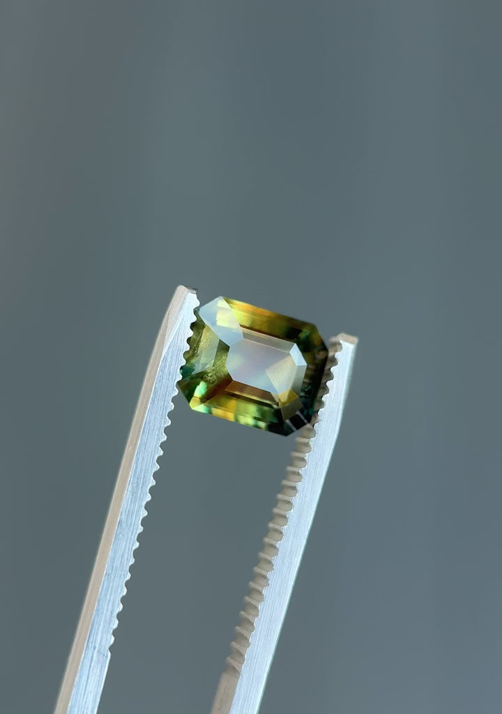 Rotating close-up video of an emerald cut sapphire held in tweezers, capturing color shifts of green, yellow, and grey under natural light – 2.02ct from Kenya.