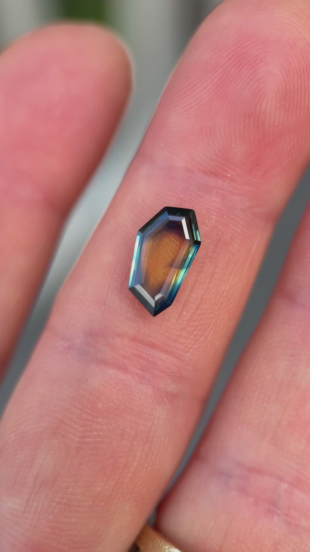 Elongated shield portrait cut sapphire in tweezers displaying natural color shifts and precision faceting.