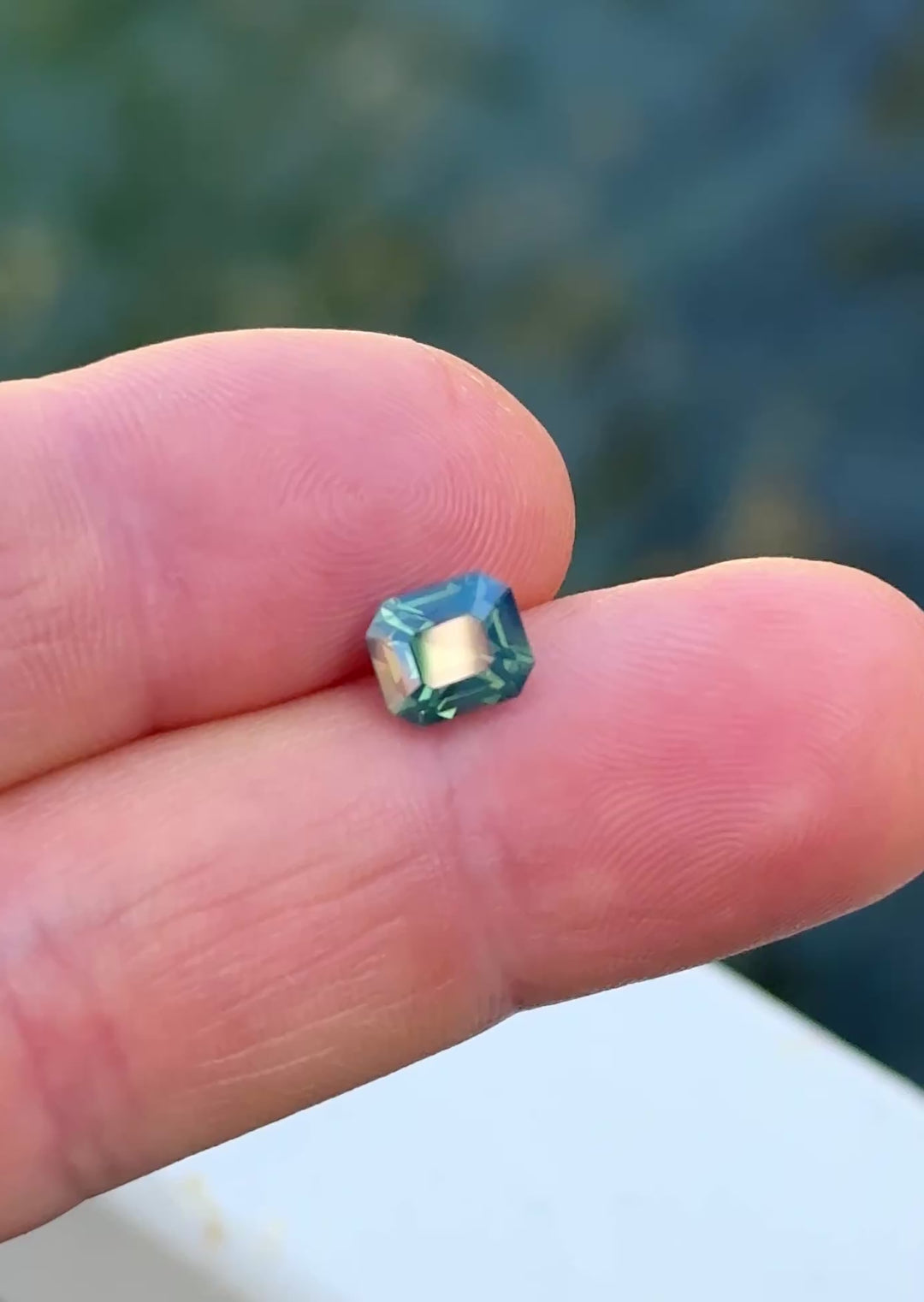 Teal opalescent precision emerald cut sapphire shown on fingertips moving from shade to direct sunlight to highlight color and sparkle.