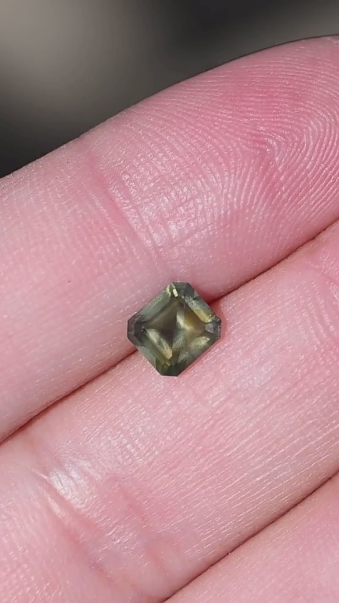 Natural 1.08 carat opalescent moss green sapphire from Gombe, Nigeria, displayed on the hand for scale. This untreated gemstone features a precision octagonal step mix cut, muted olive-green coloration and distinctive internal texture. An unusual loose sapphire for custom rings and bespoke fine jewelry.