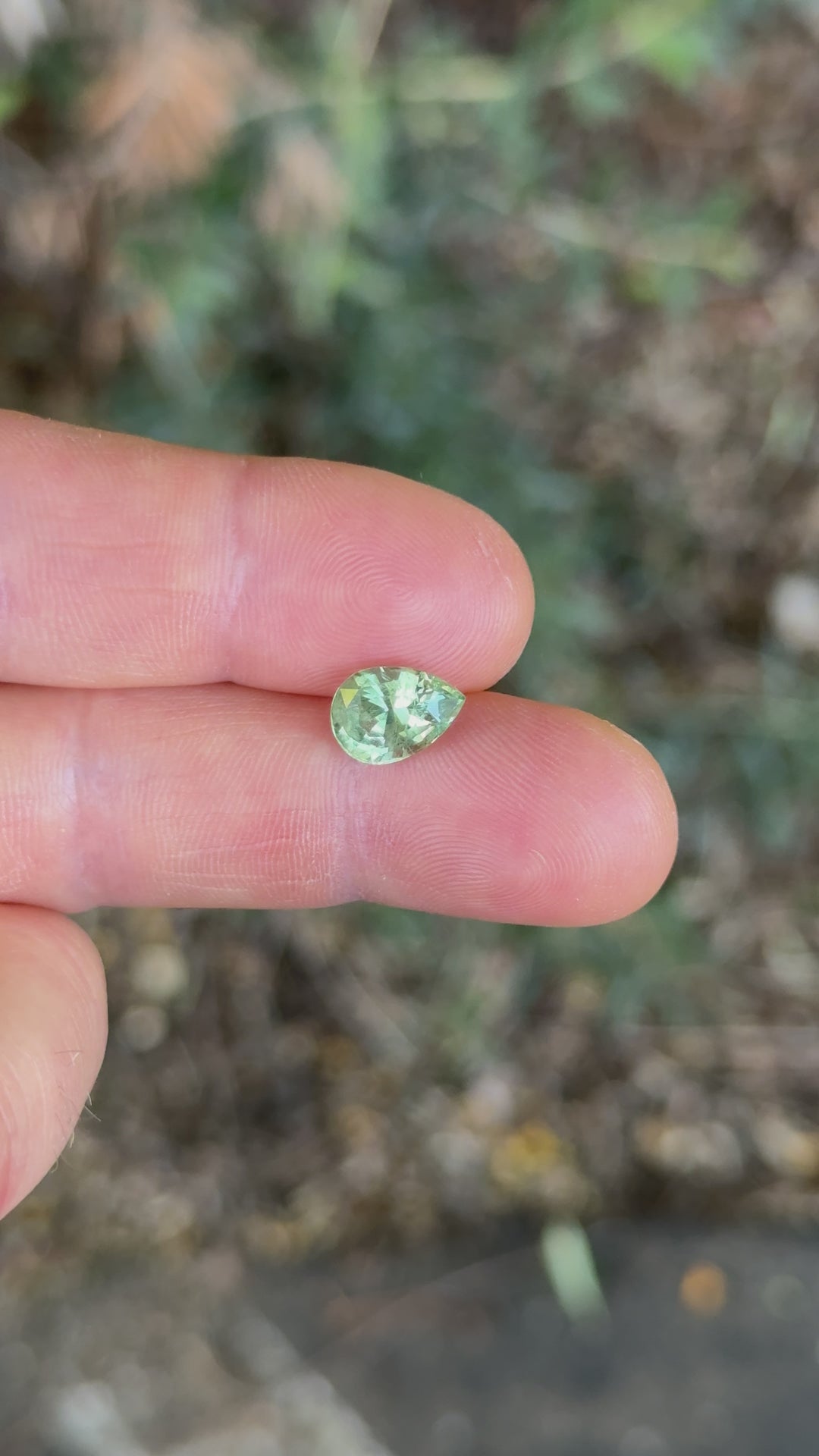 Rotating view of a 3.04ct pear-shaped sage green and peach sapphire held in fingers under natural daylight, showcasing its brilliance and color play